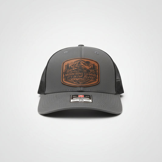 Gray trucker hat featuring a leather ‘Mountains and Mud, It’s In Our Blood’ patch on a light gray background, Hollarwood Outfitters