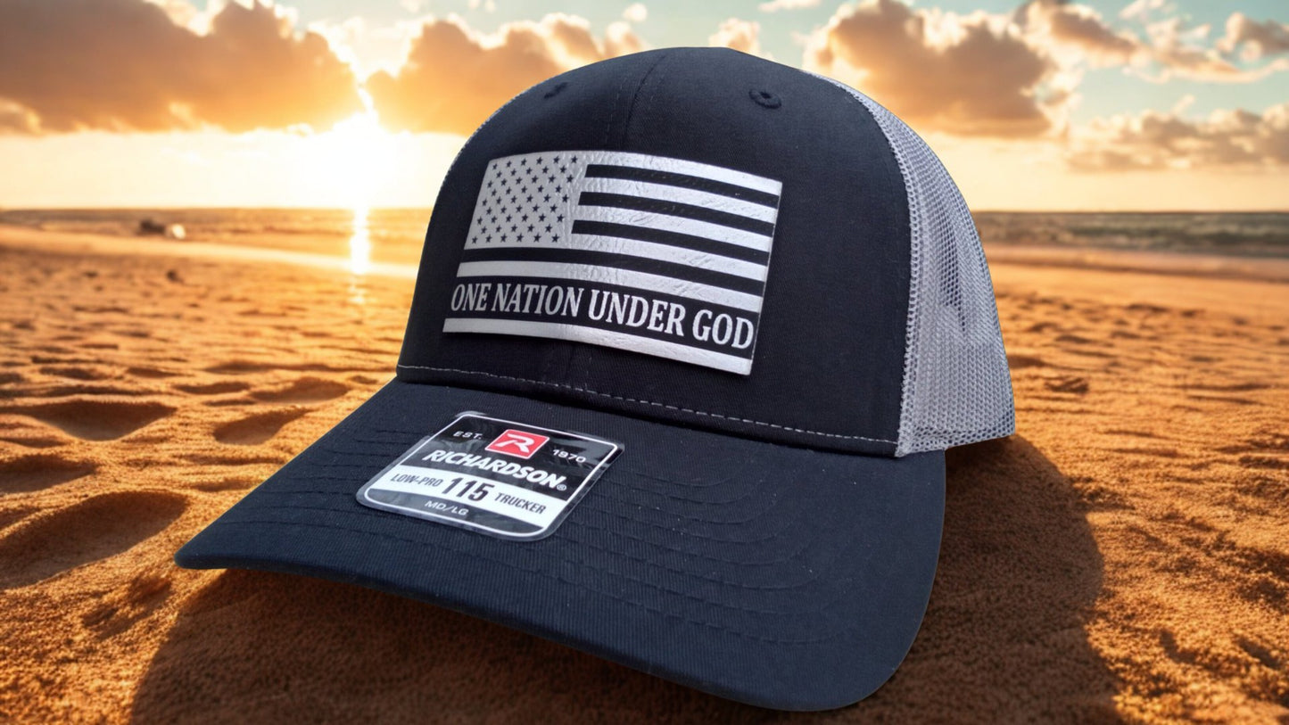 Black and charcoal trucker hat featuring a leather American flag ‘One Nation Under God’ patch on a sandy beach at sunset, Hollarwood Outfitters