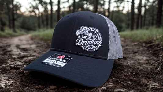 Black and gray trucker hat featuring a leather ‘Day Drinking Club’ patch on a dirt path with trees in the background, Hollarwood Outfitters